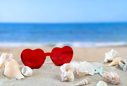 Stock photo showing close-up view of red, heart-shaped sunglasses besides a pile of seashells on a sunny, golden sandy beach with sea at low tide in the background.