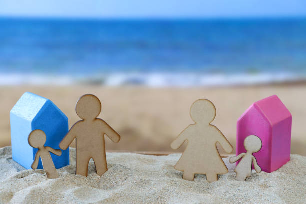 Close-up image of wood cutout separated family, blue and pink wooden model holiday cottages on sand, parents and children, father and son, mother and daughter, sandy beach background with waves breaking at low tide, sea coastline, divorced family holiday stock photo