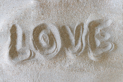 Stock photo showing close-up, elevated view of writing drawn on sunny beach with word 'love' written in soft golden sand on seaside coastline.