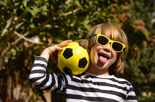 Smiling little boy, 5 years old, holding a yellow soccer ball. A child in sunglasses, a summer day, enjoys the holidays, plays in kindergarten