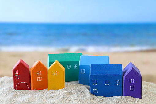 Stock photo showing close-up view of group of rainbow coloured model coastal houses in red, orange, yellow, green, light and dark blue, and purple on a sand pile on a sunny beach with the sea and clear blue sky in the background. Real estate, vacation home and beach hut rental concept.