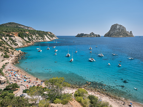 Wide-angle view of the bay of Cala d'Hort, located on the southern coast of Ibiza, renowned for its crystal-clear waters and its scenic view on the iconic rocky isles of Es Vedrà and Es Vedranell. The dazzling bright light of a Mediterranean summer noon, a perfectly clear sky, sailing boats and yachts rocking gently on the water, colourful cliffs covered with lush pine trees and a wide variety of Mediterranean shrubs and plants, tourists engaged in many different activities. High level of detail, natural rendition, realistic feel. Developed from RAW.