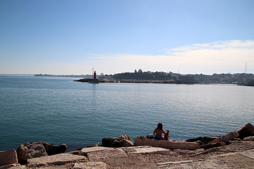 Trani, Province of Barletta-Andria-Trani, Italy - 04 february 2024: the pier of Sant'Antonio with the red lighthouse, and a sailing boat leaves the port. In the background, the fort and the historic public garden.