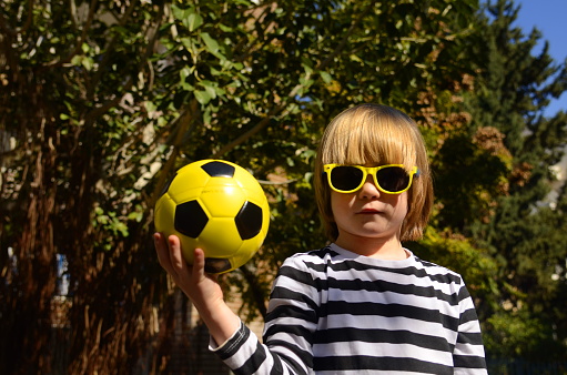 Cute little preschooler boy holding a yellow soccer ball. A child in sunglasses holds a ball, laughs