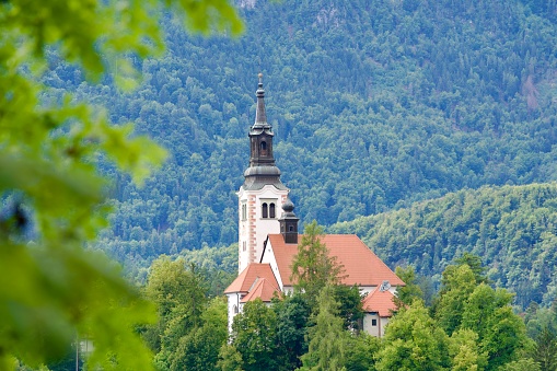 Lake Bled with close-up of church on an island and woodland in the background on a cloudy summer day. Photo taken August 8th, 2023, Bled, Slovenia.