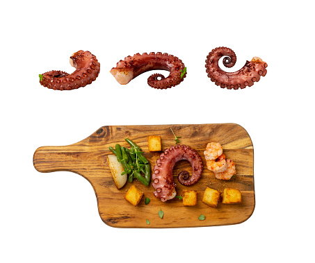 Grilled octopus tentacle on a wooden cut board served with shrimps, green beans, burnt onion and potatoes isolated with clipping path. Delicious barbecue seafood dish with fresh greens top view