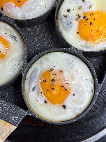 Stock photo showing close-up view of four fried eggs that are being cooked in a stainless steel, non-stick frying pan on a ceramic hob. The pan has rings to crack eggs into in order to keep them separate and stop the egg whites from merging.