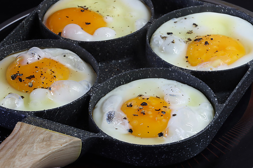 Stock photo showing close-up view of four fried eggs that are being cooked in a stainless steel, non-stick frying pan on a ceramic hob. The pan has rings to crack eggs into in order to keep them separate and stop the egg whites from merging.