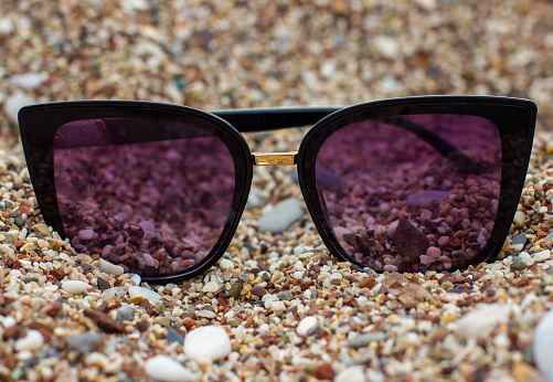 Stock photo showing close-up view of sandy beach with a pair of octagonal, metal framed, tinted, mirrored sunglasses on damp sand at sea water's edge at low tide, against a backdrop of woodland with palm trees.