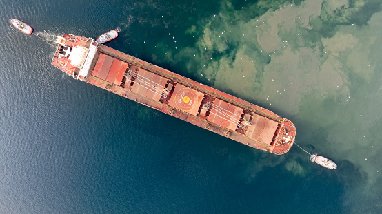 Aerial top down view of Tug boats assisting big cargo ship. Large cargo ship enters the port escorted by tugboats.