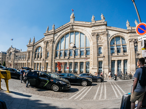 Paris, France - May 20, 2018: ultra wide-angle view of French street with iconic tall building of Gare du Nord signage large crowd of people commuting in the Pre-Covid era - taxis, cars cobblestone floor