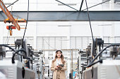 in large industrial plants There are machinery to produce steel and plastic for export. It is owned and operated by a female Asian engineer, holding a laptop, wearing a suit, hard hat, and radio.