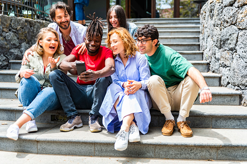 Portrait of cheerful multiracial young friends watching at smart phone - Mixed race people sitting stairs using mobile phone - young friends sitting outdoors and looking at mobile phone