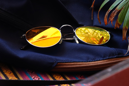 Stock photo showing close-up view of a pair of tinted, mirrored sunglasses resting on a navy blue backpack on tablecloth covered table.