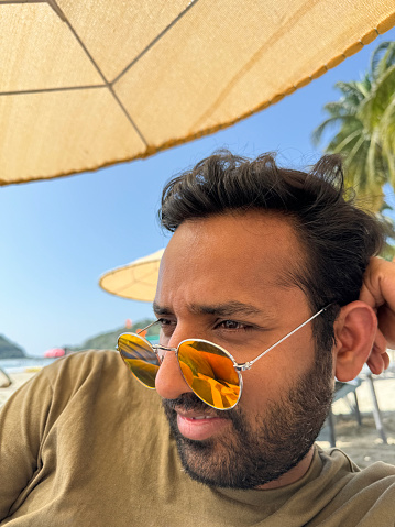 Stock photo showing close-up view of Indian man lying under beach umbrella posing in yellow tinted mirrored sunglasses whilst leaning on elbow.