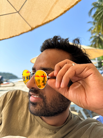 Stock photo showing close-up view of Indian man lying under beach umbrella posing in yellow tinted mirrored sunglasses whilst leaning on elbow.