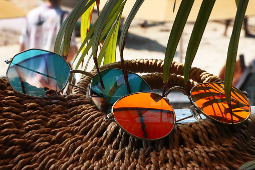 Stock photo showing close-up view of two pairs of tinted, mirrored sunglasses resting on a seagrass handbag, beach umbrellas and sun parasols on golden sandy beach with sea in the background.