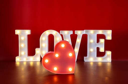 Stock photo showing close-up view of pink, heart-shaped box lined with mounted illuminated in front of light bulbs illuminated love sign, with the word 'LOVE' being spelt out by individual letters lit by bright white light bulbs.. Valentine's Day and romance concept.