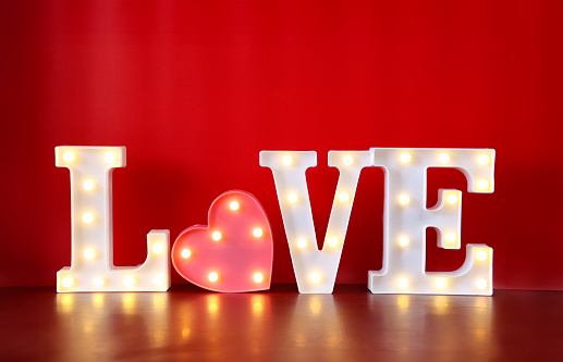 Stock photo showing an illuminated love sign, with the word 'LOVE' being spelt out by individual letters lit by bright white light bulbs. Pink, heart shaped box lined with mounted illuminated, light bulbs replacing the letter 'O'. Valentine's Day and romance concept.
