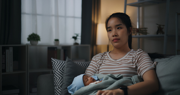 Night scene, Newborn sleeping in young mother's arms, She use remote change channel television to relaxation in living room