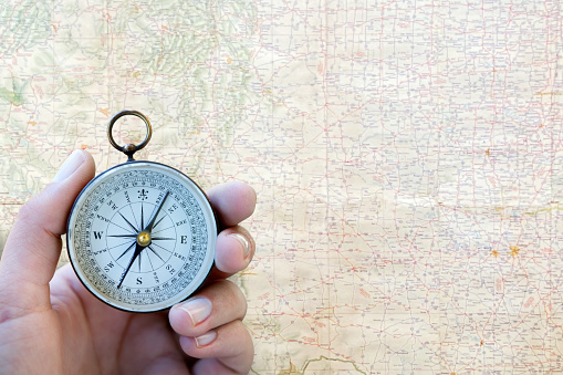 A close up of a woman's hand holding a compass with a blurred road map in the background that provides ample room for copy and text.