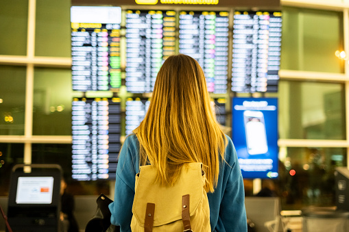 A woman stands with her back to the camera, backpack in back, checking the airport timetable leaflet. She looks attentively at the flight schedules, preparing for her upcoming journey.