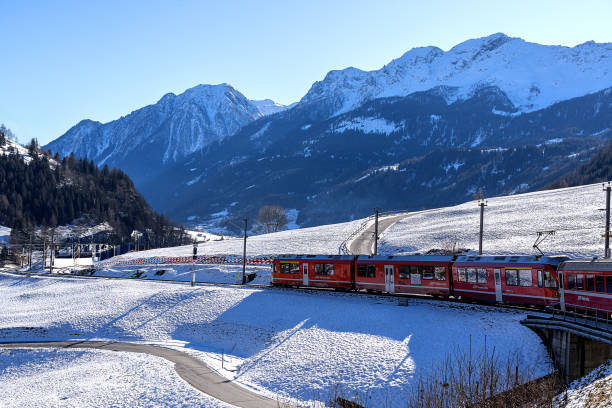 Switzerland, 01/13/2024: The UNESCO World Heritage Bernina red train slowly advances through the snow in the shadow of the conifer-covered Swiss Alps