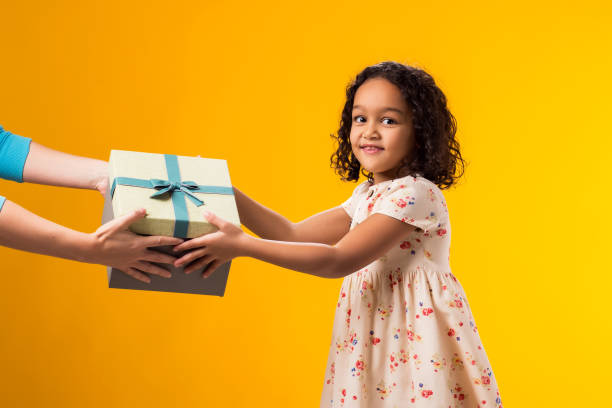 istock Smiling kid girl taking giftbox from hands over yellow background. Birthday and celebration concept 2000435405