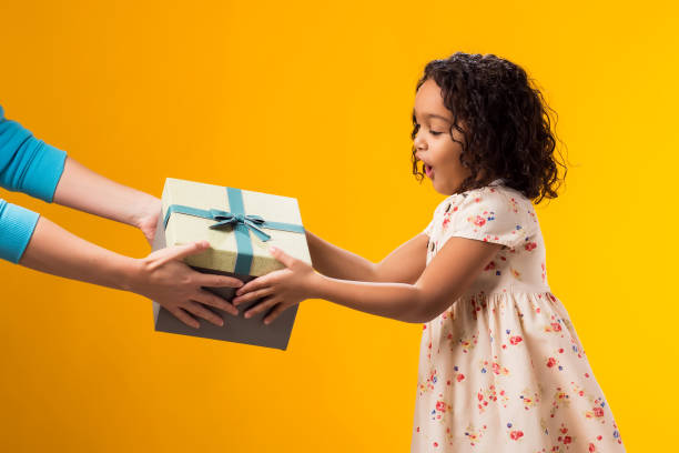 istock Smiling kid girl taking giftbox from hands over yellow background. Birthday and celebration concept 2000435406