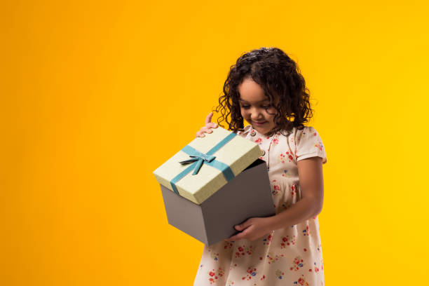 istock Smiling kid girl holding giftbox and looking inside the box over yellow background. Birthday and celebration concept 2000435408