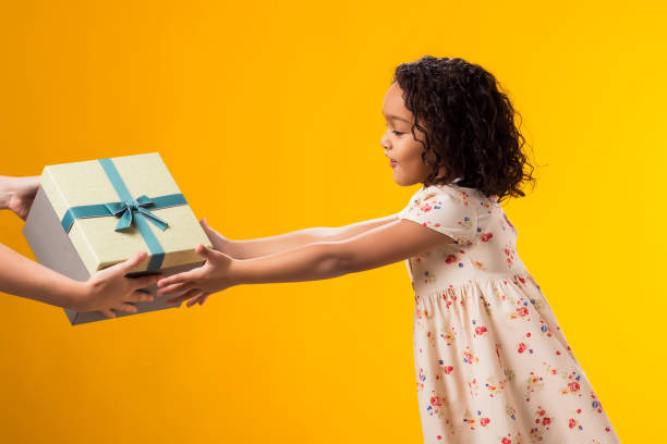 istock Smiling kid girl taking giftbox from hands over yellow background. Birthday and celebration concept 2000435410