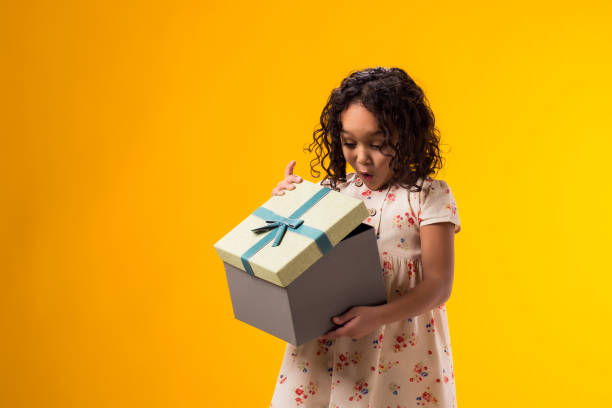 istock Smiling kid girl holding giftbox and looking inside the box over yellow background. Birthday and celebration concept 2000435412
