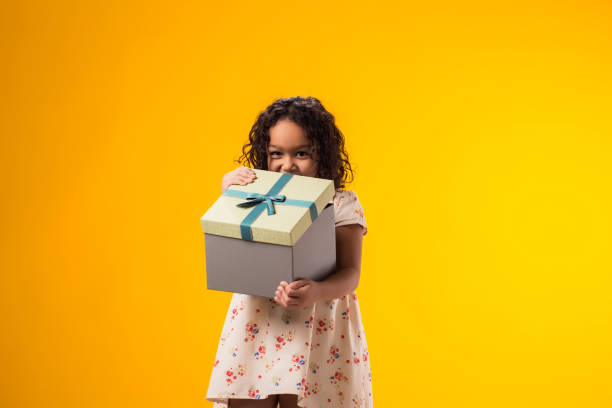 istock Smiling kid girl holding giftbox and looking inside the box over yellow background. Birthday and celebration concept 2000435414