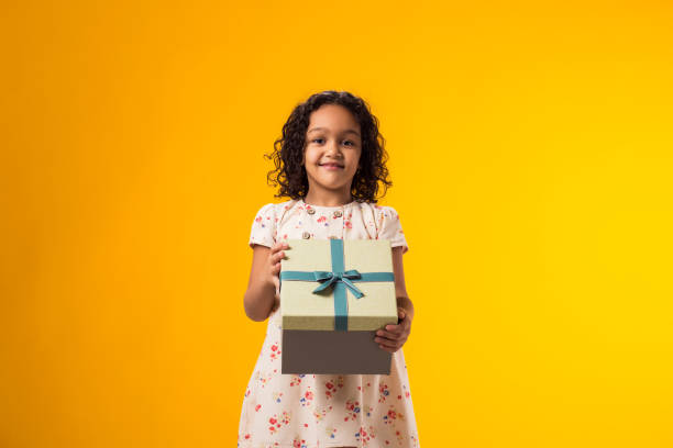 istock Smiling kid girl holding giftbox over yellow background. Birthday and celebration concept 2000435422