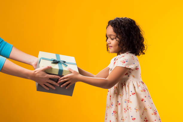 istock Smiling kid girl taking giftbox from hands over yellow background. Birthday and celebration concept 2000435443