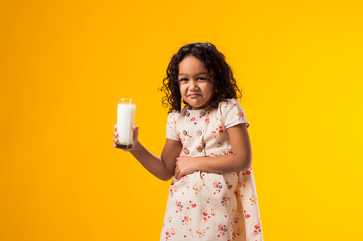 Portrait of kid girl holding glass of milk and feeling abdominal pain on yellow background. Lactose intolerance concept