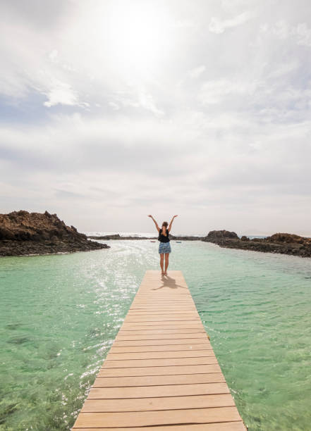 Experience pure joy as a woman stands on a wooden boardwalk, gazing out at the ocean with arms raised in happiness. The warm sunlight bathes the scene, creating a serene atmosphere. Allow the image to evoke feelings of freedom, gratitude, and the simple joy found in the embrace of the sea breez