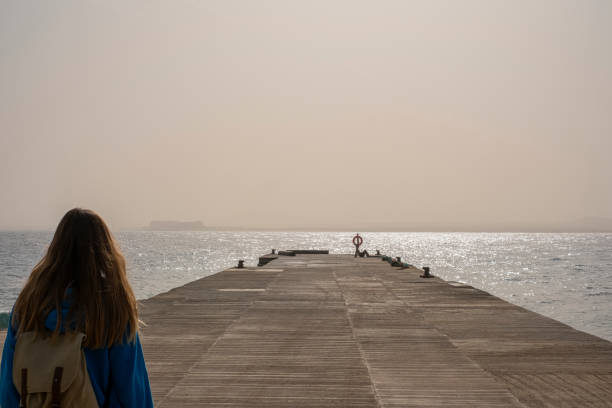 A backpacker woman embarks on a journey towards the walkway along the serene seaside. With a backpack on her shoulders, she explores the coastal path, embracing the beauty of the sea and the tranquility of the surroundings