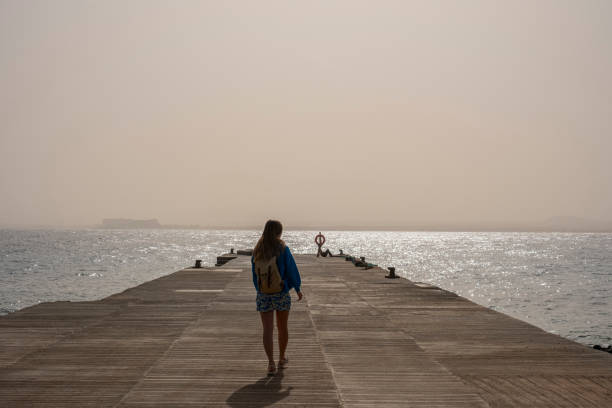 A backpacker woman embarks on a journey towards the walkway along the serene seaside. With a backpack on her shoulders, she explores the coastal path, embracing the beauty of the sea and the tranquility of the surroundings