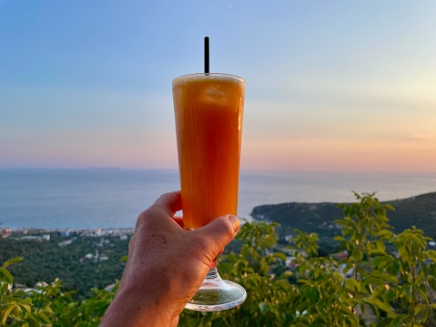 Hand holding freshly squeezed orange juice, Ionian Coast at sunset, Albania, in the background.