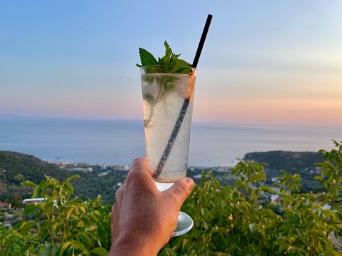 Hand holding freshly squeezed lemon juice, Ionian Coast at sunset, Albania, in the background.