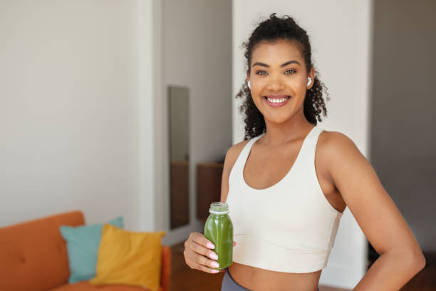 Young sporty lady with earbuds holds vegetable smoothie bottle, smiling to camera standing at home interior, enjoying healthy detox meal and playlist for domestic workout, free space