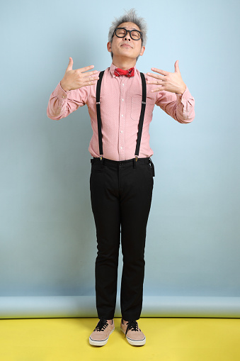 Asian senior man in black suspenders with red bow with gesture of smelling good isolated on blue background. St Valentine's Day