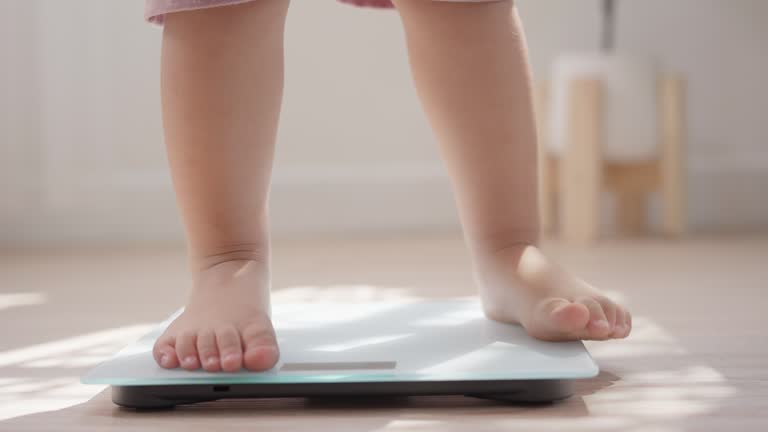 Little baby barefoot standing on weight scales on wooden floor home. Children weight control concept.