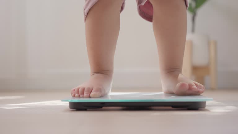 Little baby barefoot standing on weight scales on wooden floor home. Children weight control concept.