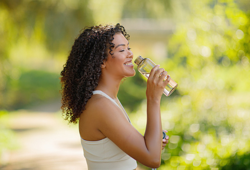 Brazilian fitness lady enjoying water drinking from bottle outdoors, posing happily in sportswear, ready for morning training in nature. Health and sport motivation. Copy space for wellness ad