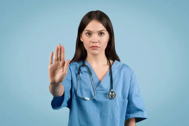 Dedicated young European lady nurse in blue medical uniform signals stop with confidence against blue background, conveying healthcare message