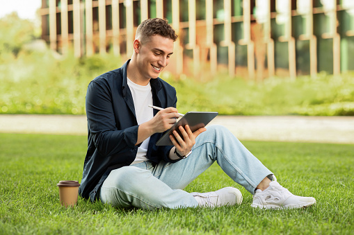 Happy millennial european man enjoy lifestyle, work on tablet with coffee cup takeaway, sit on grass in city, outdoor. Business and study at spare time near building office