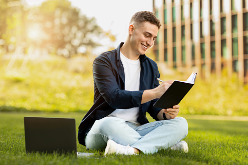 Smiling millennial european man enjoy lifestyle, work on laptop make writes a book, planning business, sit on grass in city, outdoor. Study, education, startup with technology