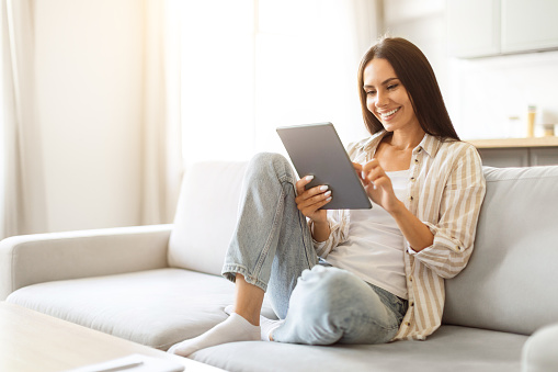 Smiling beautiful woman relaxing with digital tablet on couch at home, happy young female using modern gadget while resting in cozy living room, enjoying weekend pastime, copy space